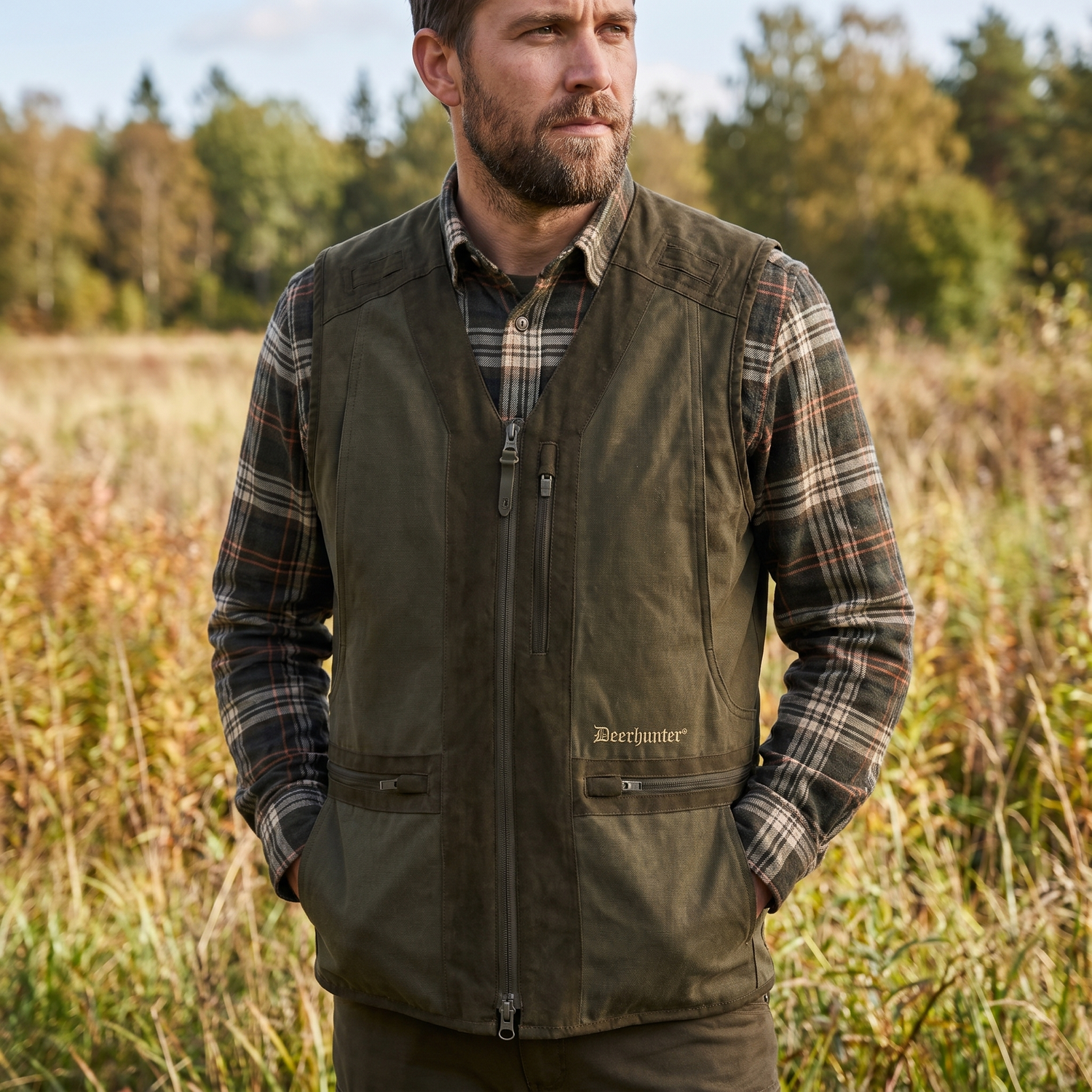 Man wearing a green vest and plaid shirt standing in a field with trees in the background