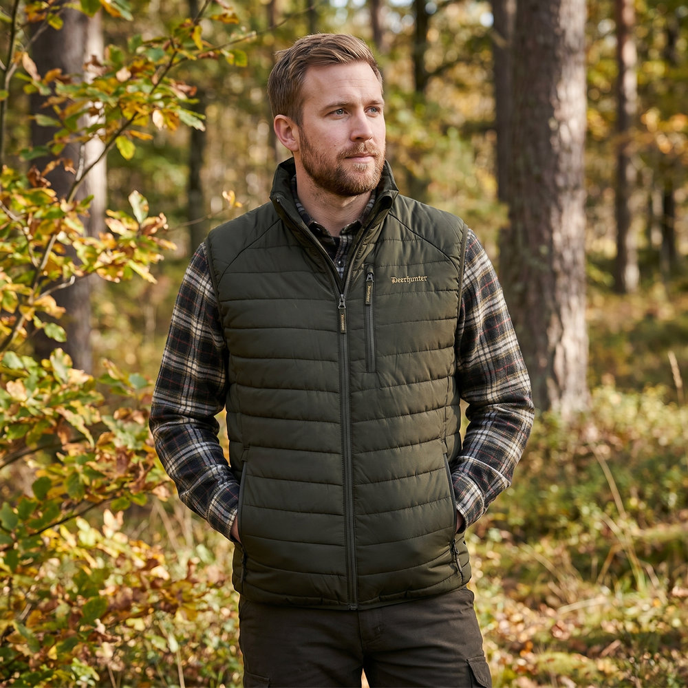 Man wearing a green puffer vest and plaid shirt in a forest setting