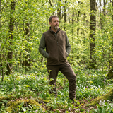 Man standing in a forest wearing green and brown outdoor clothing.